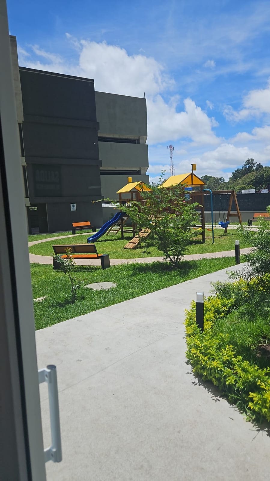 A playground area is visible, featuring a slide and climbing structure under a bright yellow canopy. Surrounding green grass and walkways provide a vibrant setting. Benches are strategically placed for relaxation, and the tall building structure offers a backdrop to this outdoor space.