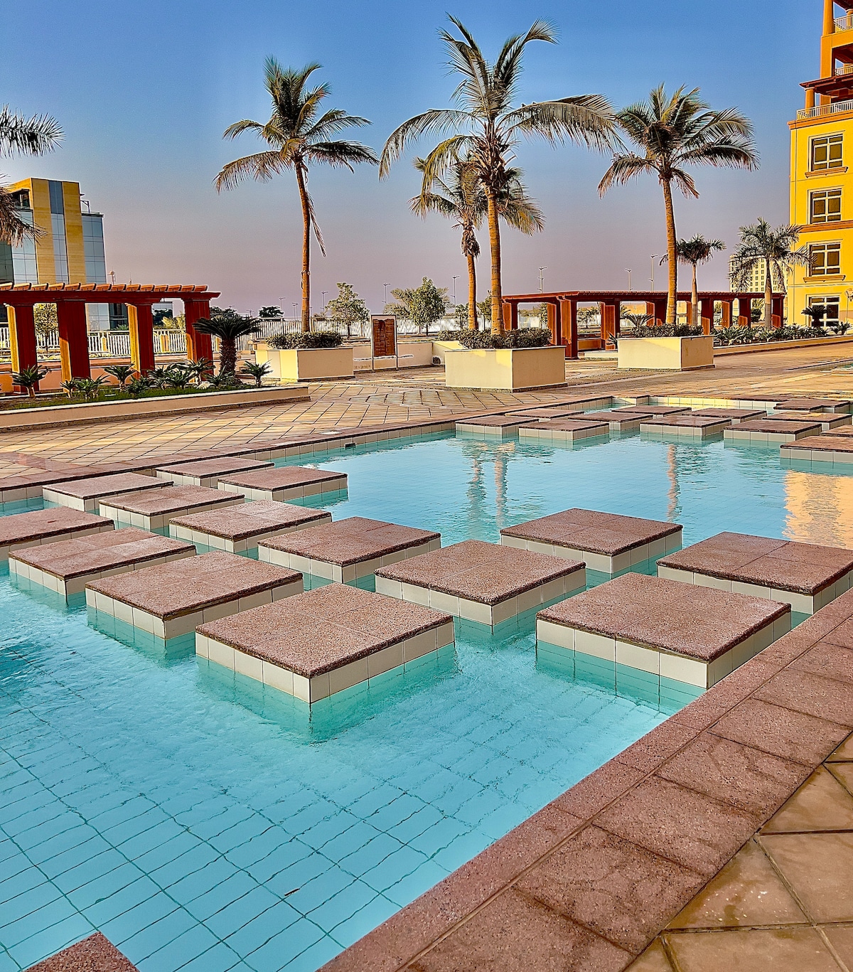 A series of stone stepping stones is set within a clear pool, creating a unique pathway across the water. Tall palm trees are visible in the background, providing a tropical ambiance. The surrounding area features paved walkways and landscaped gardens.