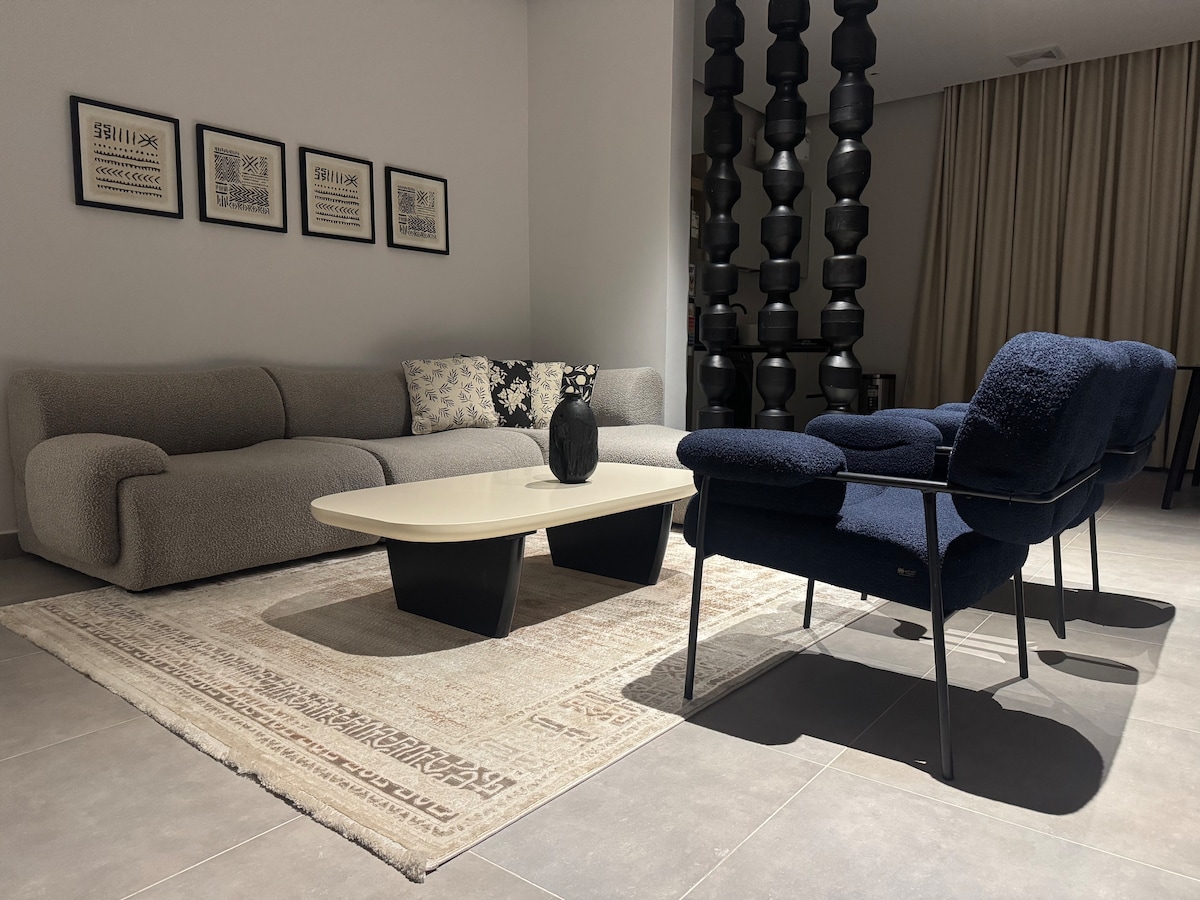 A spacious living area features a curved sectional sofa in soft gray fabric, complemented by a black decorative vase on the central coffee table. A textured area rug anchors the seating, while elegant lighting fixtures rise in the background, enhancing the overall contemporary aesthetic.