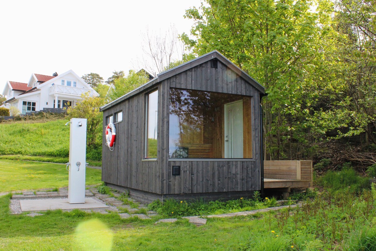 A charming wooden cabin features large windows that allow ample natural light. The exterior showcases a wooden façade with a life ring mounted on the wall. A small seating area is positioned on the deck, surrounded by greenery, offering a serene outdoor space.