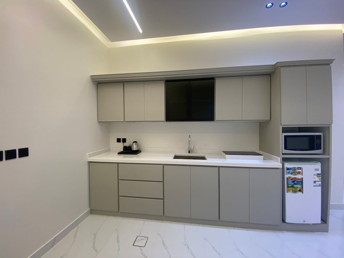 A modern kitchen features sleek, beige cabinetry complemented by a white countertop. Essential appliances, including a kettle and microwave, are discernible. A minimalist design is highlighted by subtle lighting and glossy white tiles on the floor, offering a clean and functional space.