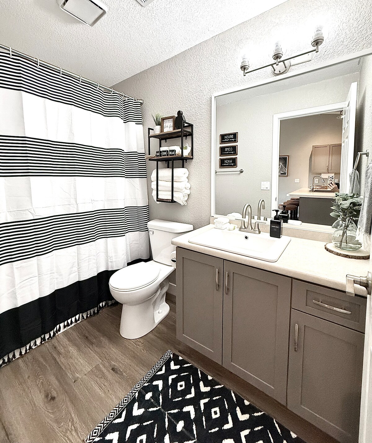 A modern bathroom is presented, featuring a clean sink area with cabinetry beneath. A black and white striped shower curtain complements the decor. A small shelf displays neatly arranged towels. The floor is covered with a geometric-patterned rug, enhancing the overall design.