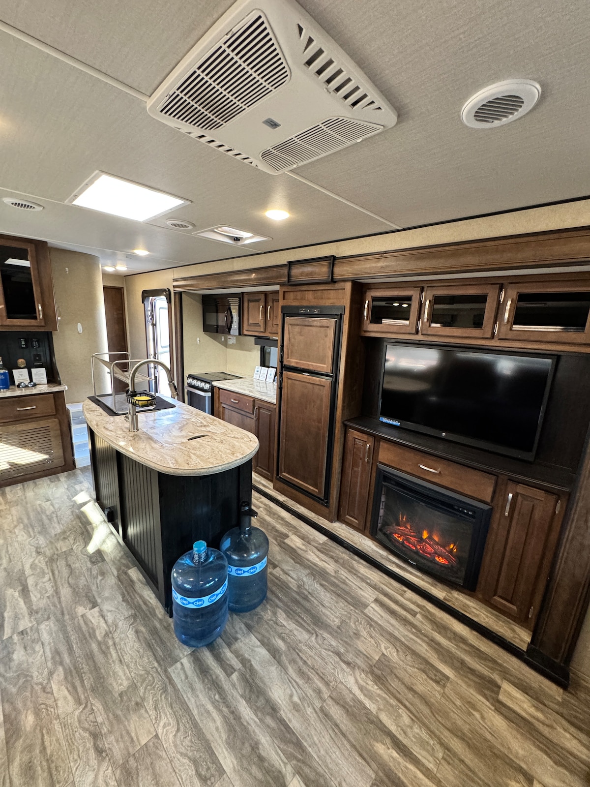 The interior of the caravan is highlighted by warm wooden cabinetry, a central kitchen island with a light-colored countertop, and modern appliances. A wall-mounted television is positioned above an electric fireplace, while two water bottles are placed near the island, enhancing the functional living space.