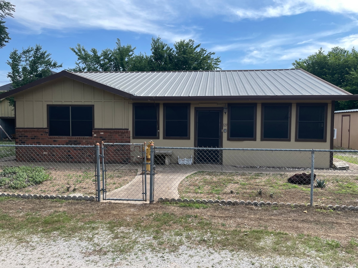 The exterior of a single-story ranch-style home features a light brown façade with brick accents. Large black-framed windows provide ample natural light. A well-maintained yard is enclosed by a chain-link fence, and a pathway leads to the entrance, flanked by sparse landscaping.