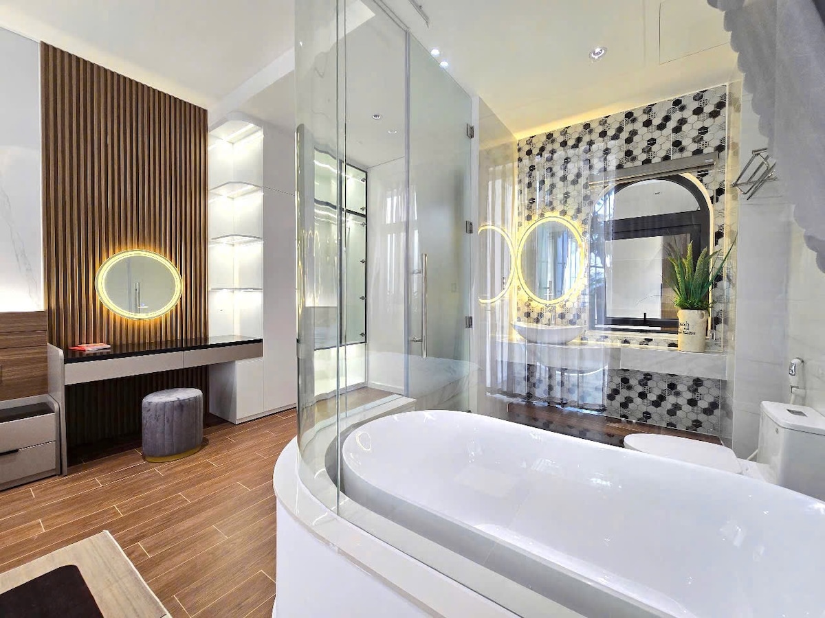 A modern bathroom is presented, featuring a sleek freestanding tub with an elegant glass partition. Ambient lighting highlights the stylish wooden accents and marble surfaces. A spacious vanity with a mirror reflects a soothing atmosphere, complemented by greenery near the tub.
