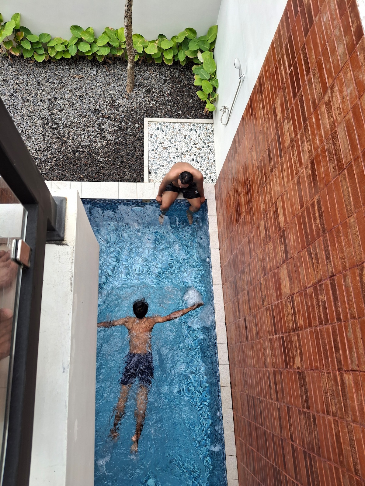 A private pool is featured with clear blue water, surrounded by smooth tiles and textured walls. A person is seen swimming while another sits at the edge, with a lush green area seen in the background, providing a tranquil setting.