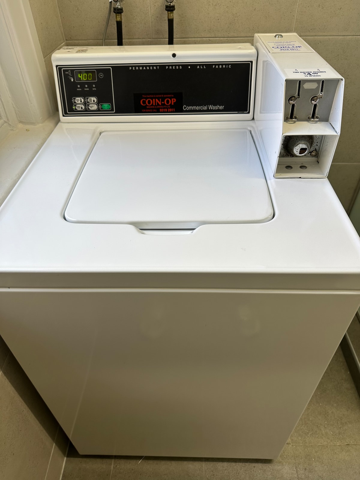 A commercial coin-operated washer is displayed, featuring a clean white exterior. The control panel is visible, showing settings and coin slots. The washer is positioned against a neutral-toned wall, providing an accessible laundry option for guests.