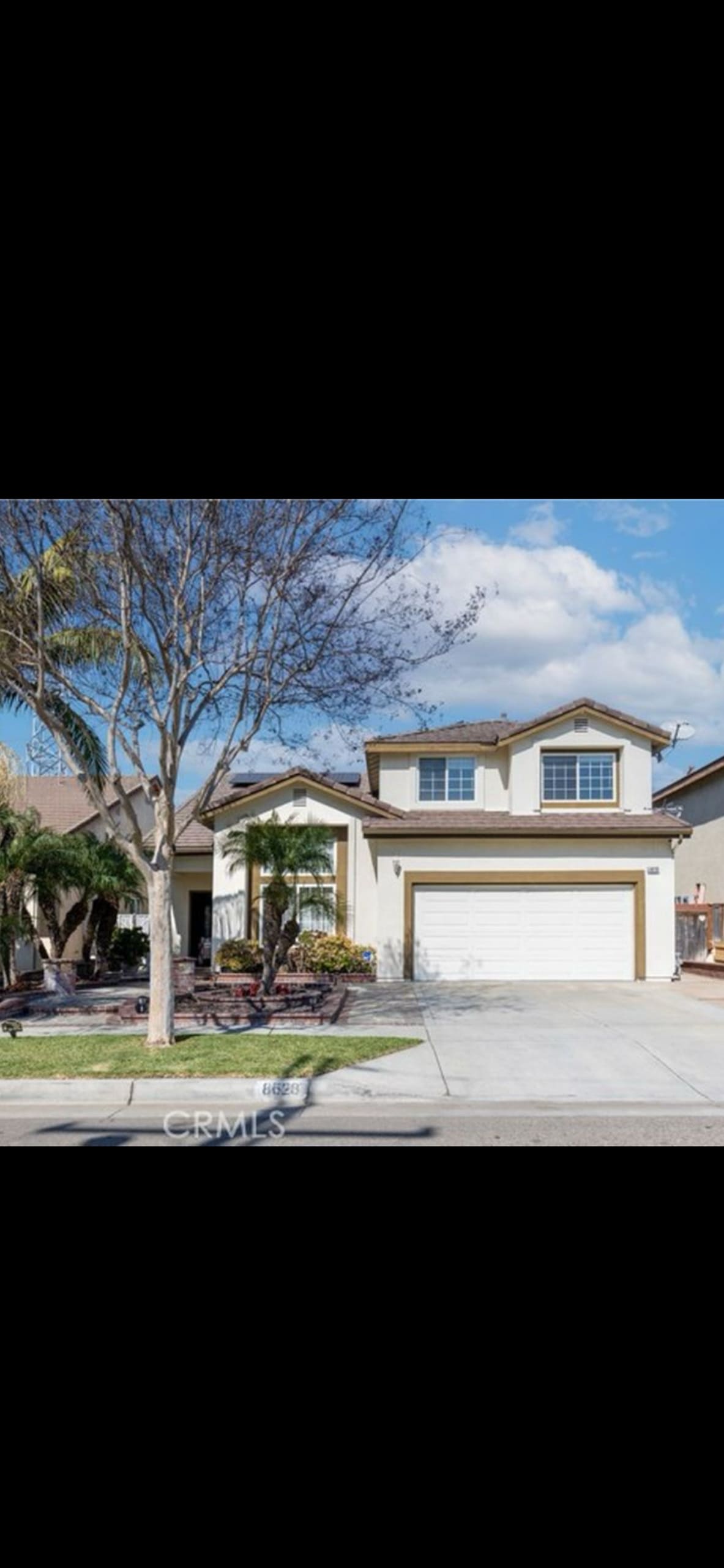 A two-story home is situated on a well-manicured lot, featuring light stucco exteriors and a double garage. A tree and lush plants are visible in the front yard, with a clear blue sky and clouds providing a serene backdrop.