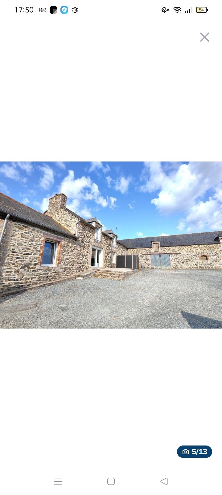 A renovated stone farmhouse is depicted, featuring a blend of rustic charm and modern elements. The structure is framed by a gravel courtyard, with several windows allowing natural light to enter. The sky above is dotted with fluffy clouds, creating a serene backdrop.