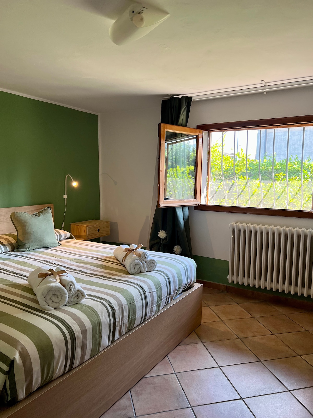 A well-lit bedroom features a spacious bed with a striped green and white duvet. Fresh towels are neatly arranged at the foot of the bed. A window allows natural light to enter, revealing views of greenery outside. A compact nightstand and radiator complete the room.