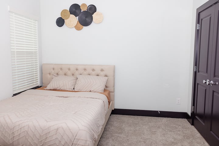 宁静的卧室，配有簇绒床头板、柔软的米色床上用品和舒适的地毯。 自然光透过百叶窗照进来，现代墙面装饰增添了一丝时尚气息。 放松、休息和充电的完美空间。