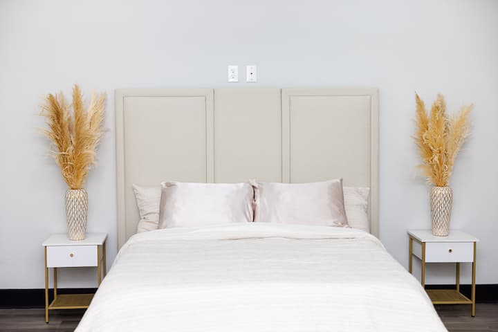 优雅简约的卧室，配有高大的软垫床头板、缎面枕头和干净的白色床上用品。 这处空间两侧放着相配的床头柜，里面的时尚花瓶中种着南美洲草，营造出宁静、高档的氛围，非常适合度过宁静的夜晚。