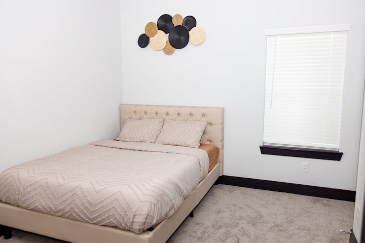 舒适的卧室，配有簇绒米色床头板、柔软的中性床上用品、毛绒地毯和现代墙面艺术品。 自然光线透过百叶窗照进来，营造出宁静而迷人的空间，非常适合放松身心。