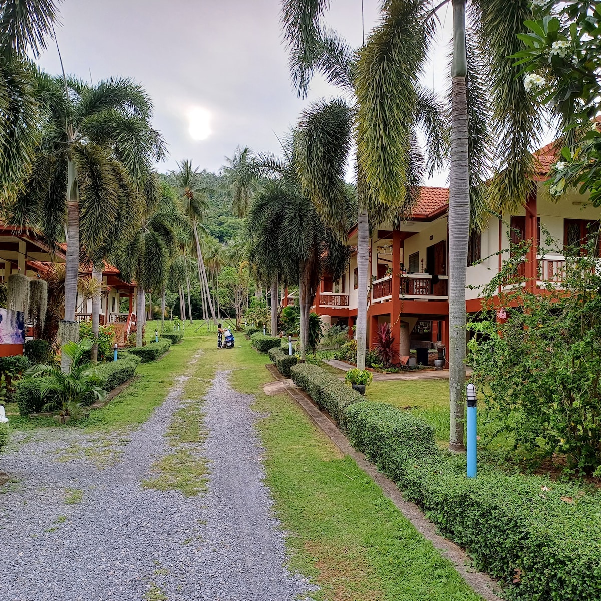 A serene pathway lined with palm trees leads to multiple two-story buildings, each featuring balconies. Well-kept gardens with hedges and green lawns are visible on either side of the path. A golf cart is parked along the gravel drive, with mountains in the background.