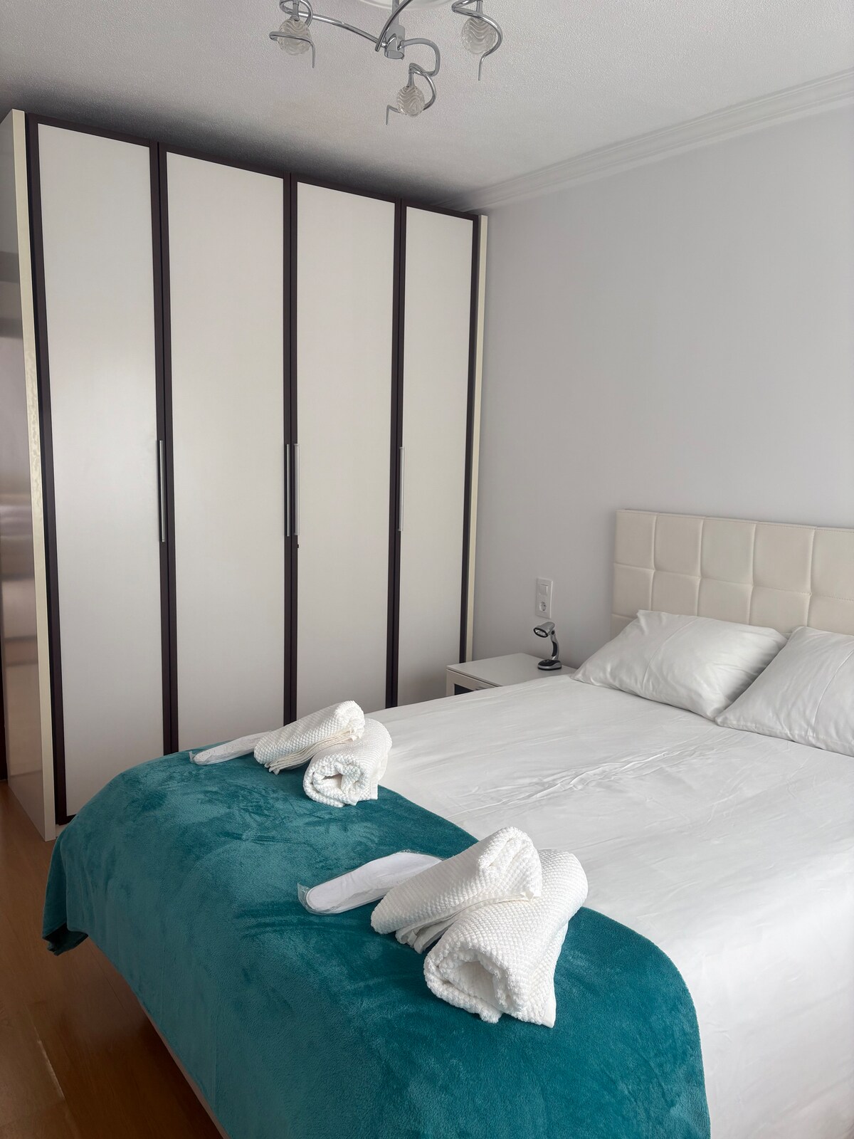 A bright bedroom features a spacious bed with a white duvet and neatly arranged towels. A modern wardrobe stands against one wall, offering ample storage. A bedside table is positioned beside the bed, contributing to the room's functional layout.