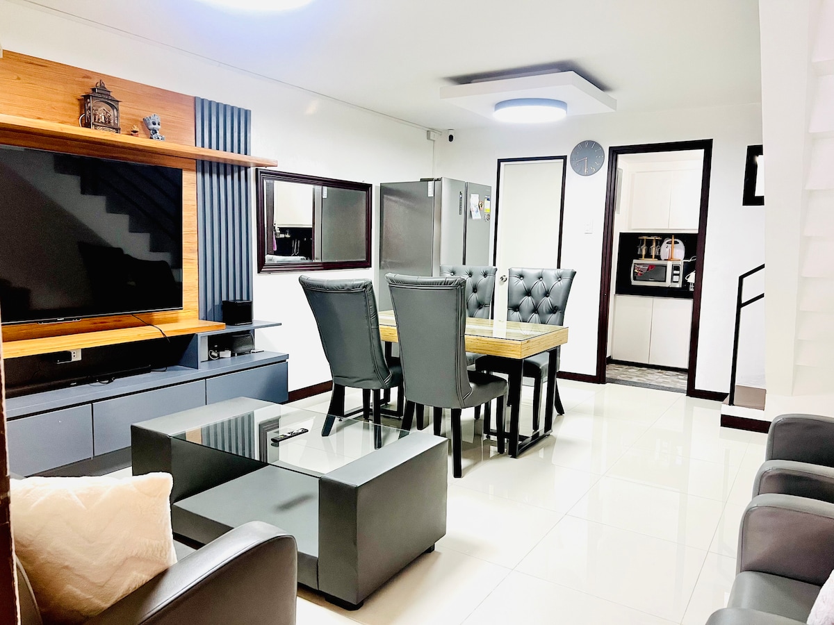 A modern living area features a wooden accent wall and a large flat-screen TV. A stylish dining table is surrounded by black upholstered chairs. A glass coffee table sits in the center. The space is well-lit with bright, white lighting.