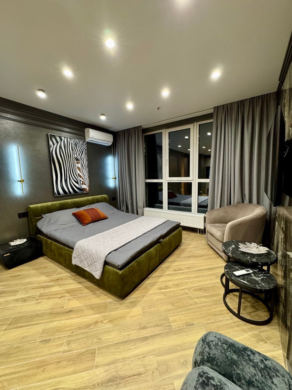 A stylish bedroom features a spacious bed with a muted green upholstered headboard, complemented by an array of decorative pillows. Natural light enters through large windows, illuminating the warm wooden floor. A comfortable armchair and small side table add to the inviting space.