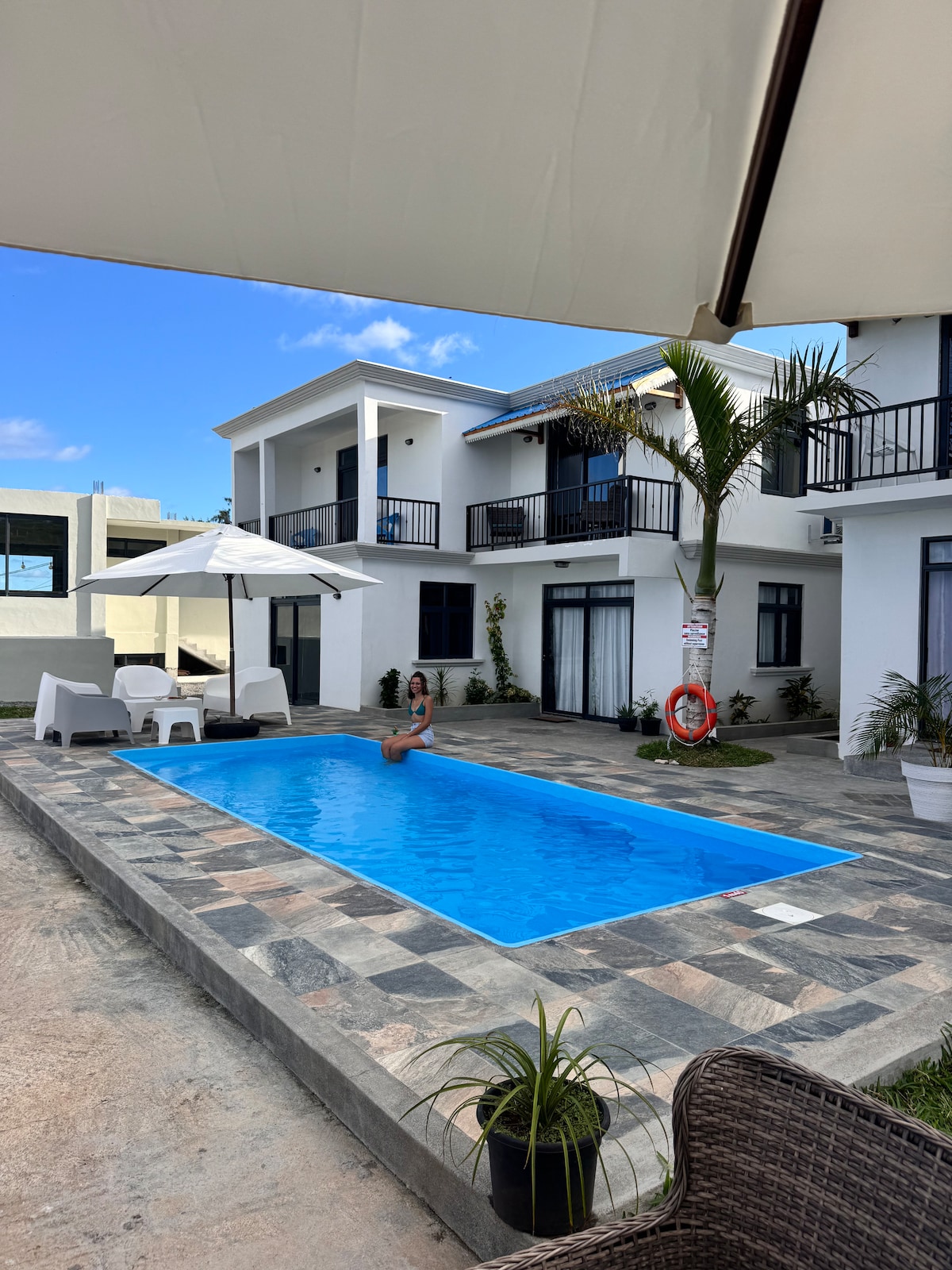A serene swimming pool is framed by modern lounge chairs and a sun umbrella. An inviting patio surrounds the pool, with a palm tree adding a touch of greenery. The contemporary two-story building is visible in the background, featuring balconies and large windows.