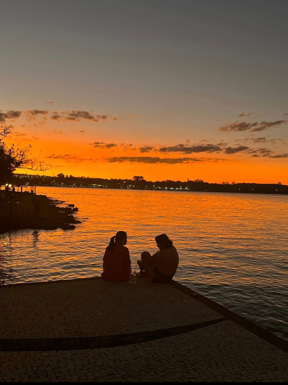 A tranquil lakeside view is captured at sunset, with vibrant orange and yellow hues reflected on the water. Two individuals are seated at the water's edge, facing each other, creating a serene and intimate setting. Silhouetted trees line the shore, enhancing the peaceful atmosphere.