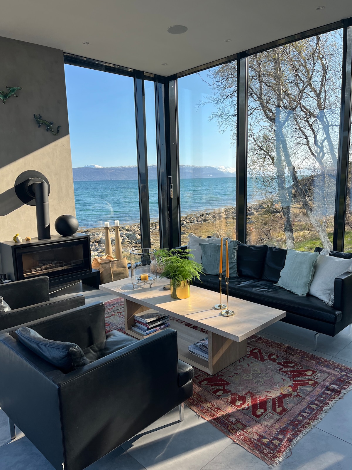 A modern living area possesses floor-to-ceiling windows that frame a stunning view of the sea. Comfortable black seating surrounds a light wooden coffee table, creating a welcoming space. Natural light floods the room, complementing the minimalist décor and enhancing the serene ambiance.