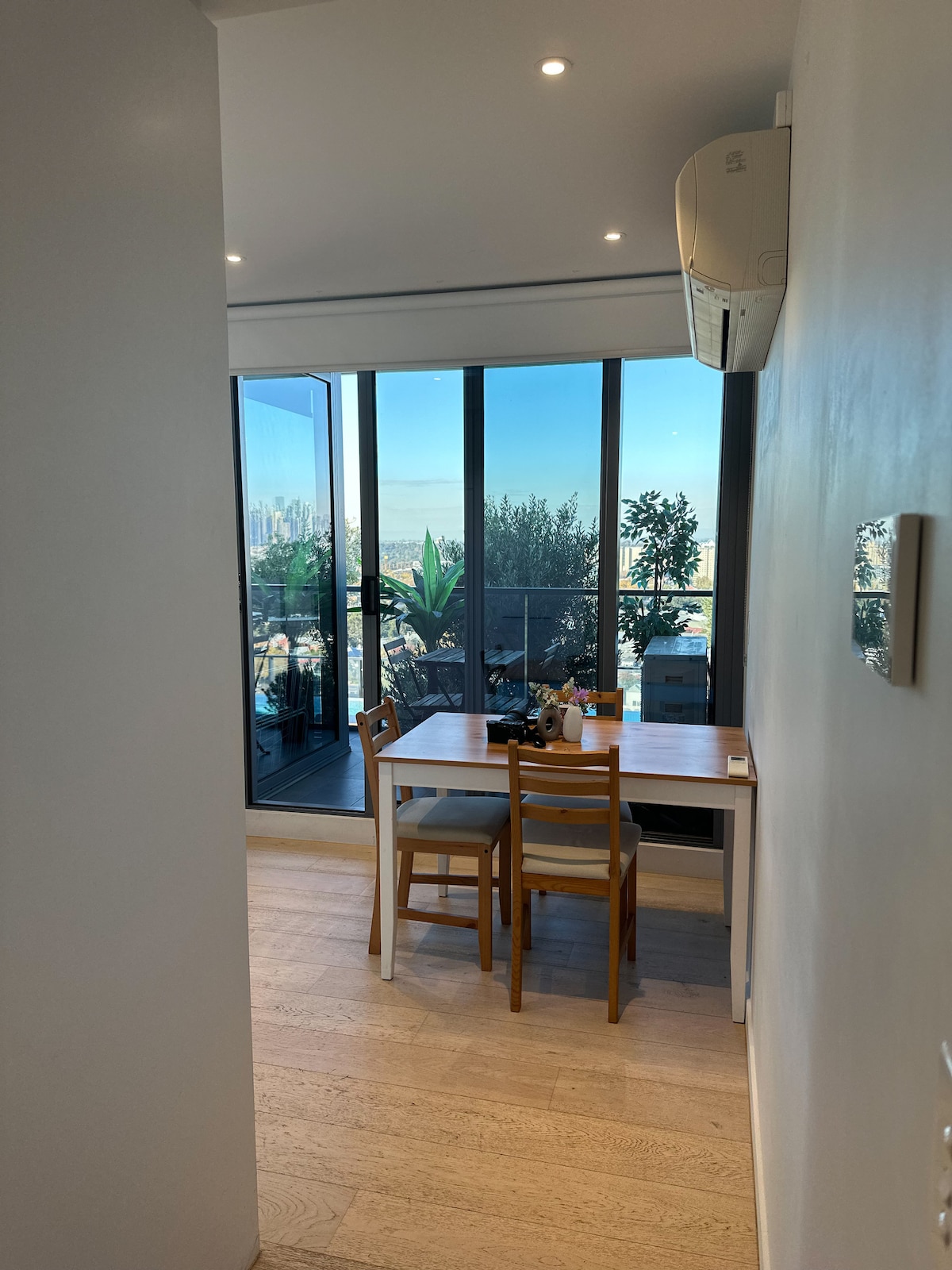 A bright dining area is seen through an entrance, featuring a small table with two wooden chairs. Floor-to-ceiling windows offer views of greenery and the skyline beyond, inviting natural light into the space.