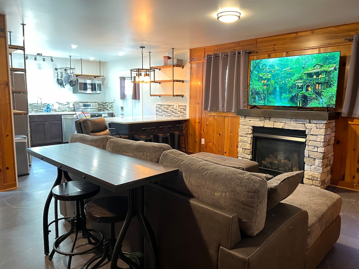 An open living area features a comfortable sectional sofa facing a stone fireplace. A large dining table is set nearby, along with modern bar stools. In the background, a well-equipped kitchen is visible, highlighted by a stylish backsplash and ample counter space.