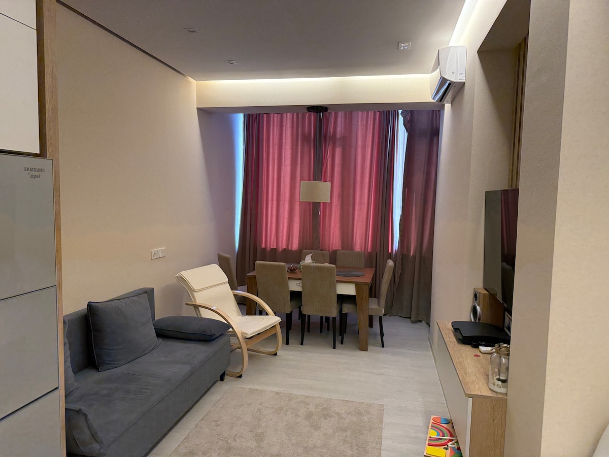 A bright living area features a comfortable gray sofa and a light-colored chair. A dining table is surrounded by four beige chairs, creating a communal space. Soft natural light enters through large, sheer curtains, adding warmth to the neutral-toned walls.