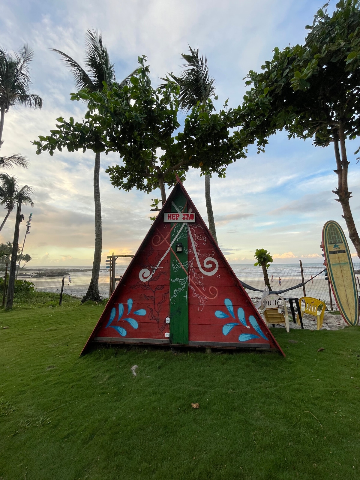 A colorful triangular cabin stands in a grassy area, surrounded by palm trees. The cabin's vibrant red exterior features artistic designs and a green door. A view of the beach and ocean is visible in the background, contributing to a tranquil outdoor setting.