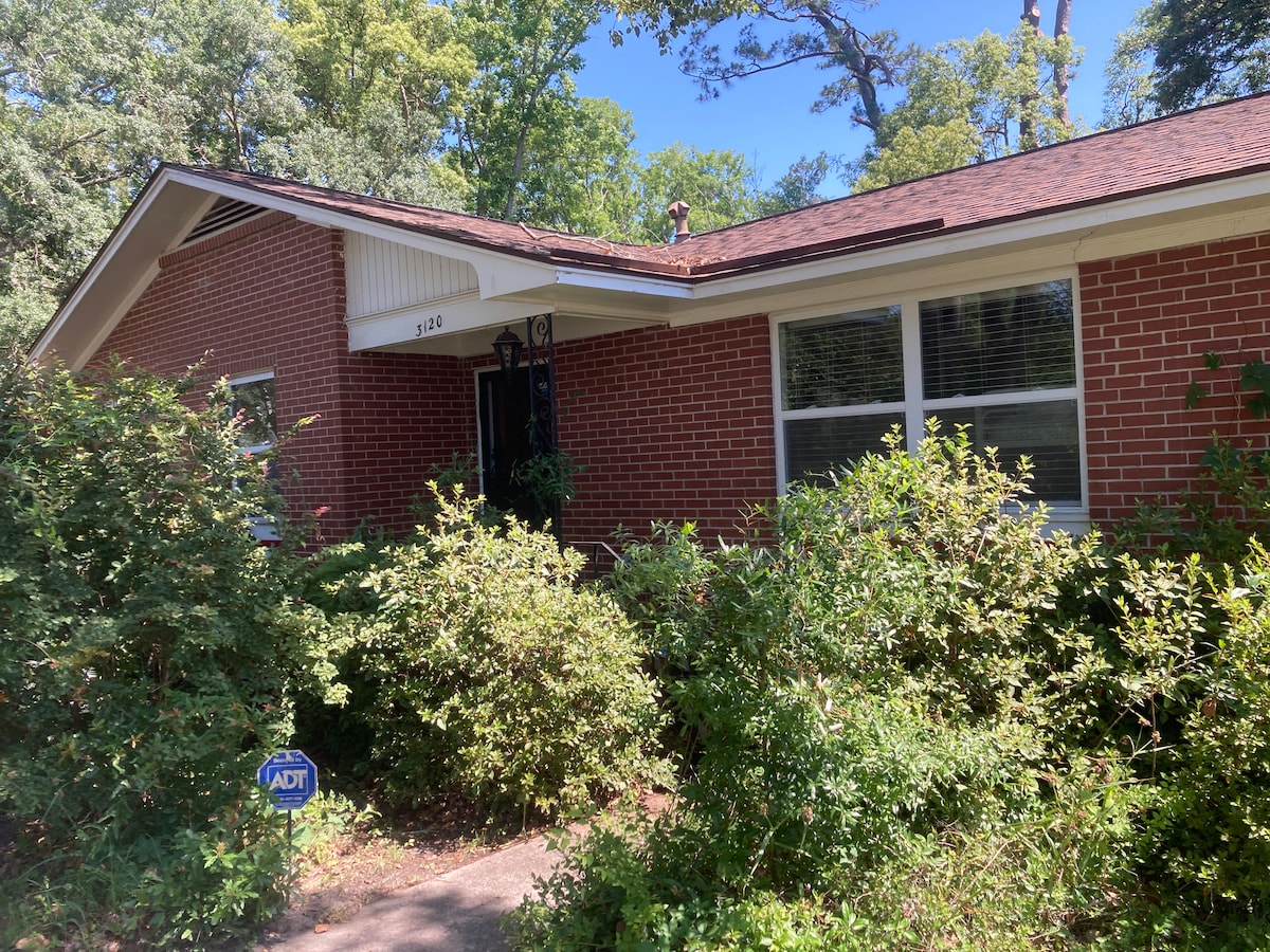 The exterior of a single-story brick home is showcased, surrounded by lush greenery and well-maintained shrubs. The entrance features a covered porch with a decorative door. Large windows allow natural light to enter, contributing to the inviting appearance of the residence.