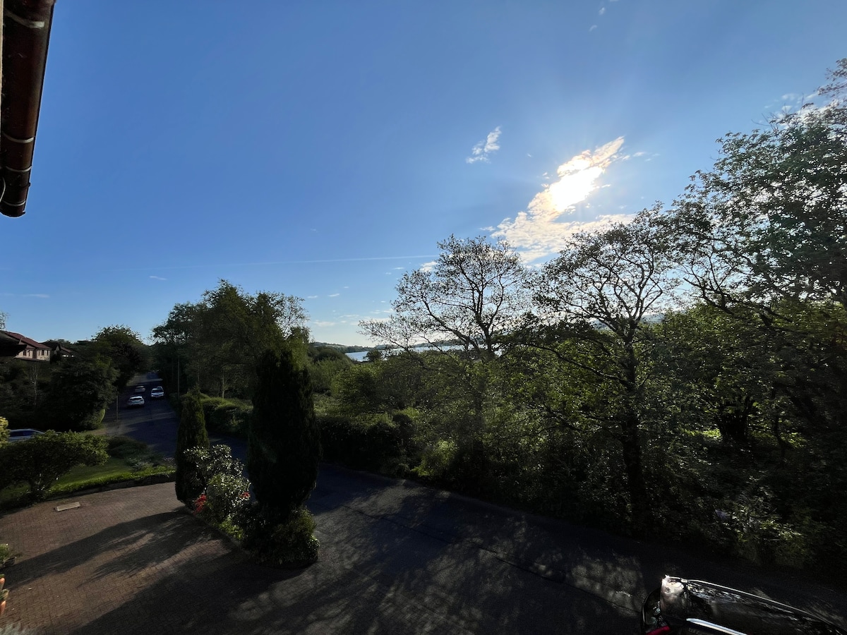 A scenic outdoor view reveals a clear blue sky adorned with a few clouds and sunlight streaming down. Lush greenery surrounds the area, with trees lining the landscape, while a glimpse of the loch can be seen in the distance.