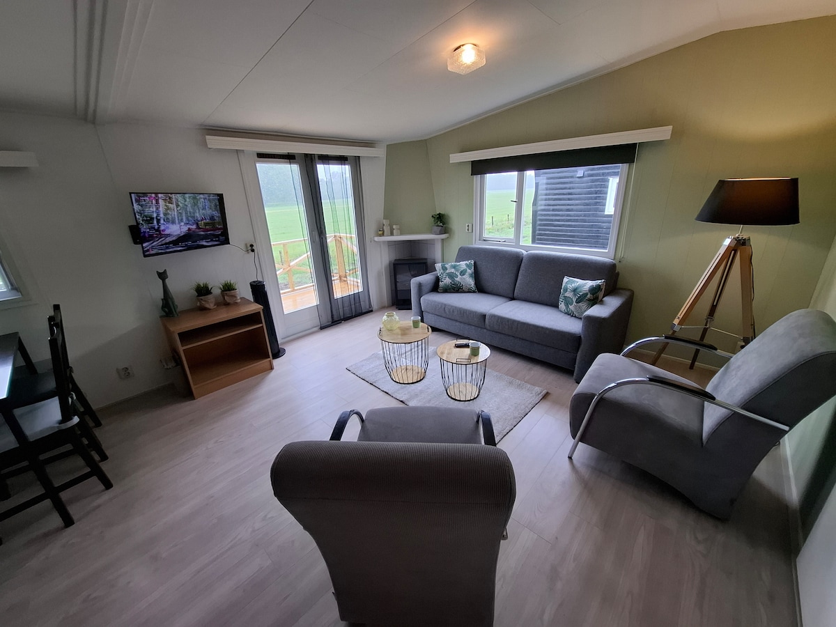 A cozy living area features a comfortable gray couch and armchair, accompanied by side tables and a coffee table. Large glass doors lead to a balcony, while a television is mounted for entertainment. Natural light brightens the space, set against a calming green wall.