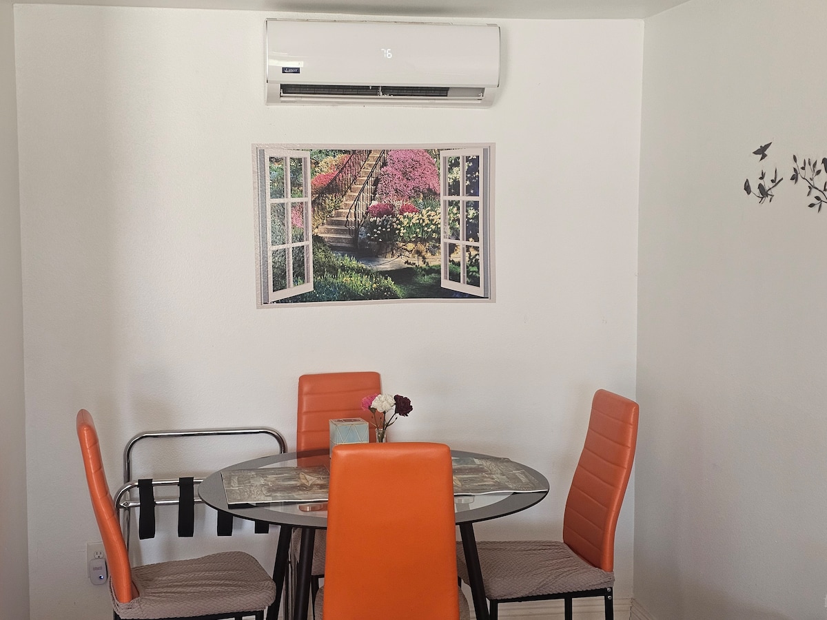 A dining area features a round glass table surrounded by four bright orange chairs. A decorative wall poster displays open windows framing a garden with colorful flowers. An air conditioning unit is mounted above, providing comfort to the space.