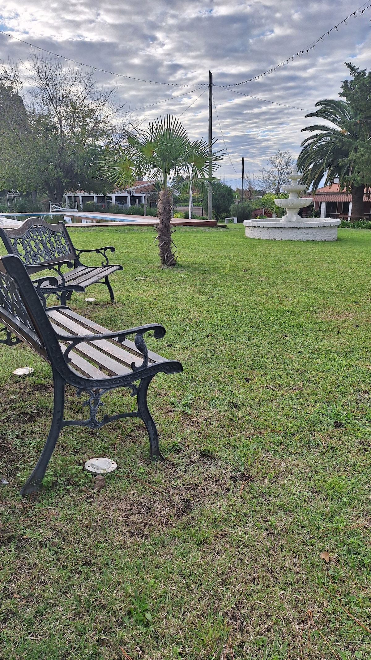A serene outdoor area is depicted, featuring several benches arranged on lush green grass. A decorative fountain stands prominently in the background, surrounded by landscaped greenery and palm trees, all under a partly cloudy sky.