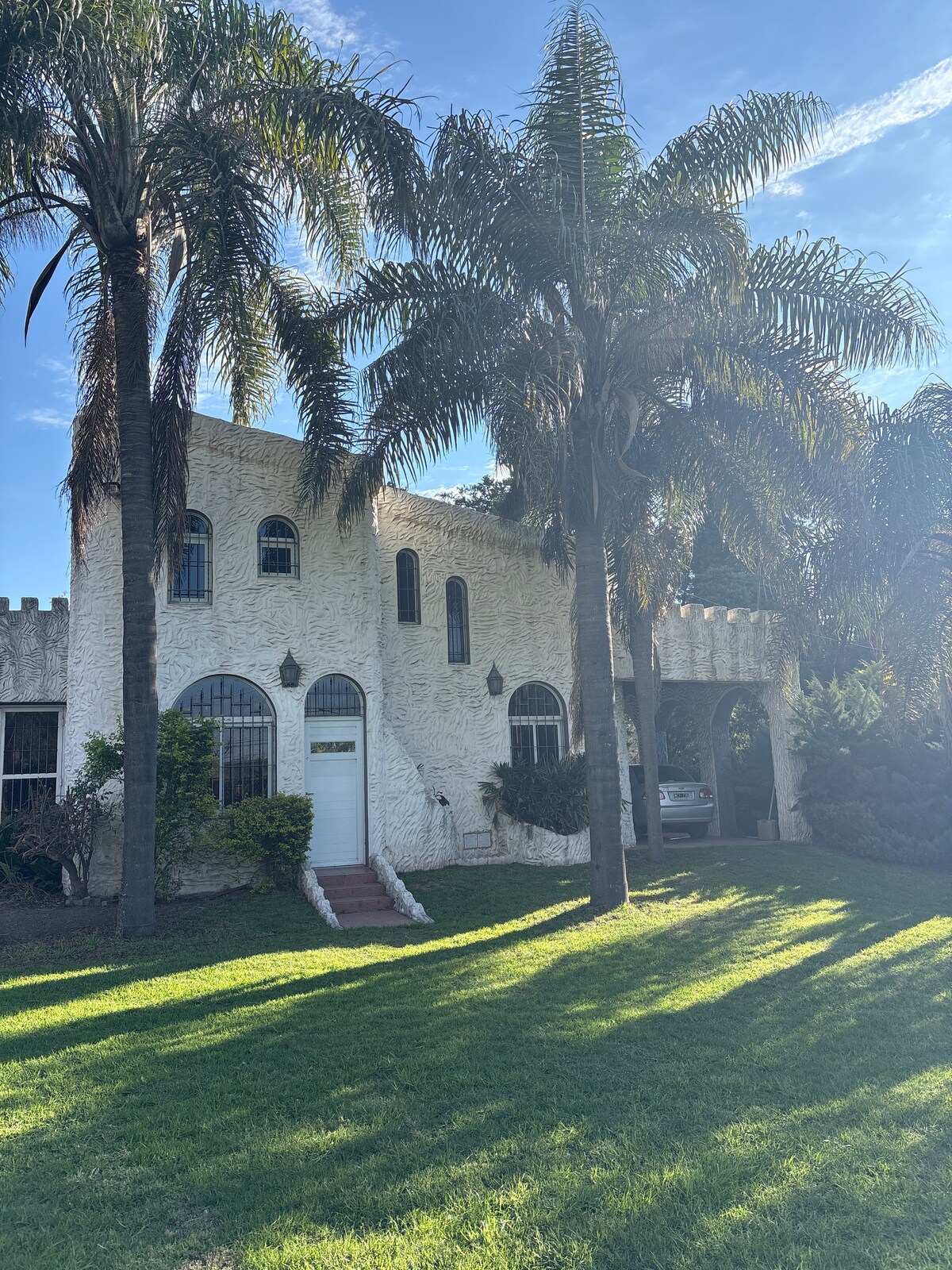 The exterior of a spacious house with castle-like features is framed by tall palm trees, creating a welcoming front yard. Large windows and a decorative entrance add character to the white facade, which is complemented by a lush green lawn under a clear blue sky.