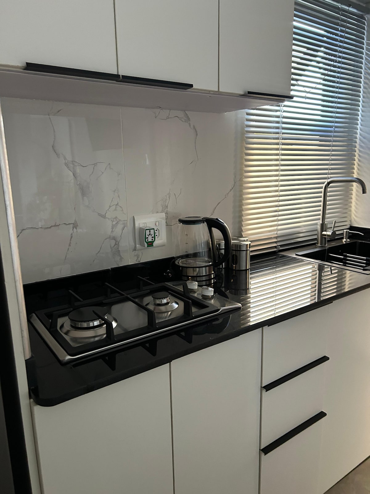 A modern kitchen area is presented with sleek black countertops and white cabinetry. A gas stove, kettle, and a coffee maker are arranged neatly, while natural light filters through the window partially covered by blinds.