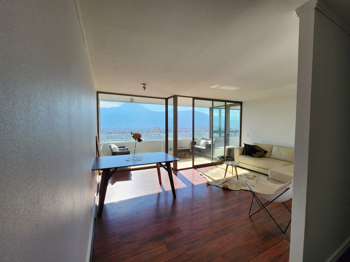 A spacious living area is illuminated by natural light, with large windows offering panoramic views of the surrounding mountains. A simple dining table is positioned beside a muted sofa, and a cozy rug adds warmth to the wooden floor. A minimalist chair complements the inviting atmosphere.
