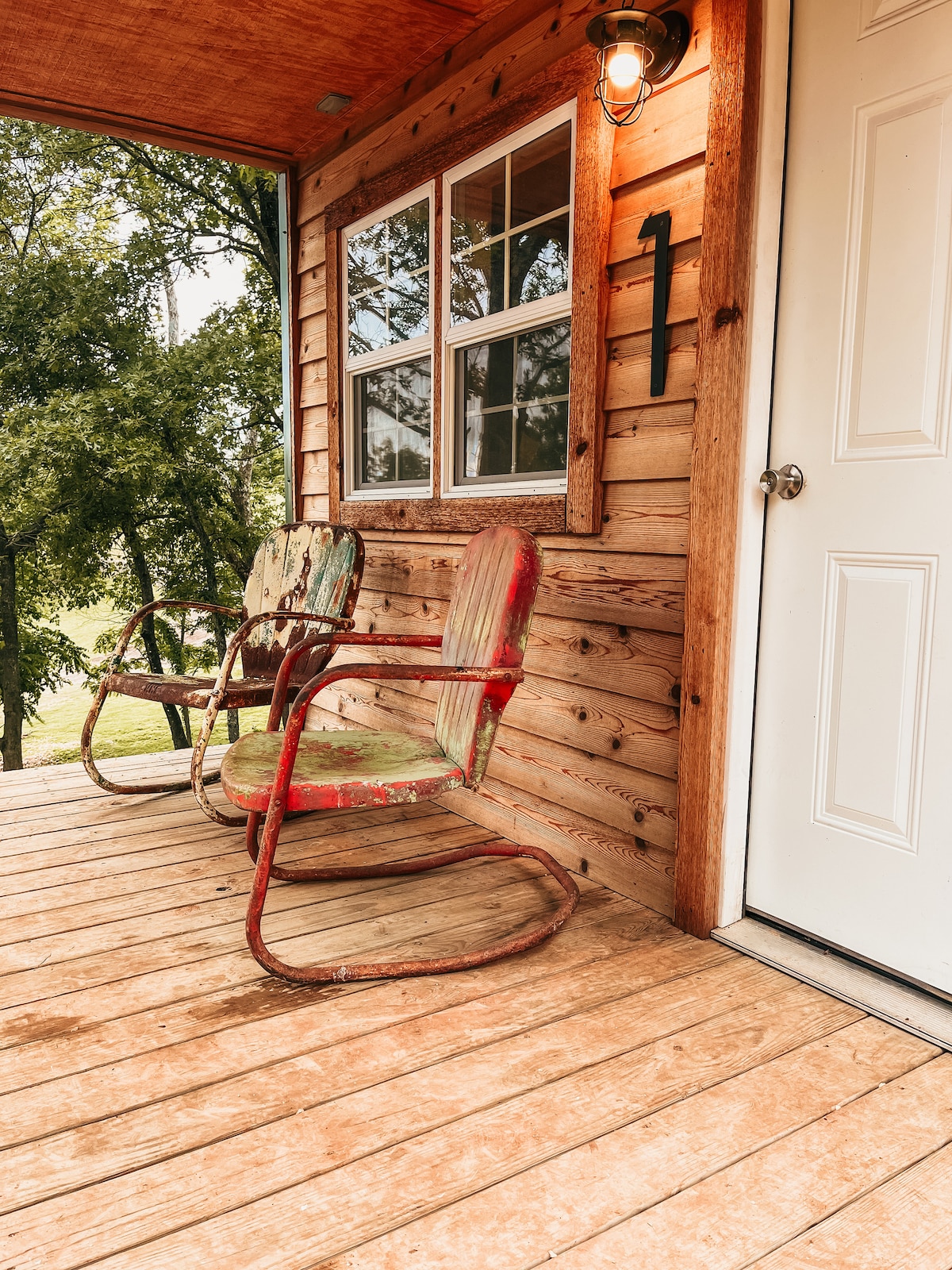 A spacious porch features two vintage rocking chairs with peeling paint, positioned on a wooden deck. Natural light streams in through a large window with four panes, framed by rustic wooden siding. A white door with a simple design is located to the right.