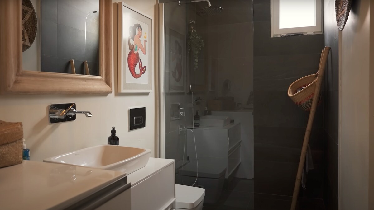 The bathroom features a modern design with a shower area enclosed by glass panels. A wall-mounted mirror is framed in wood above a sleek countertop sink. A colorful art print adorns the wall, while a woven basket and additional storage options are visible.