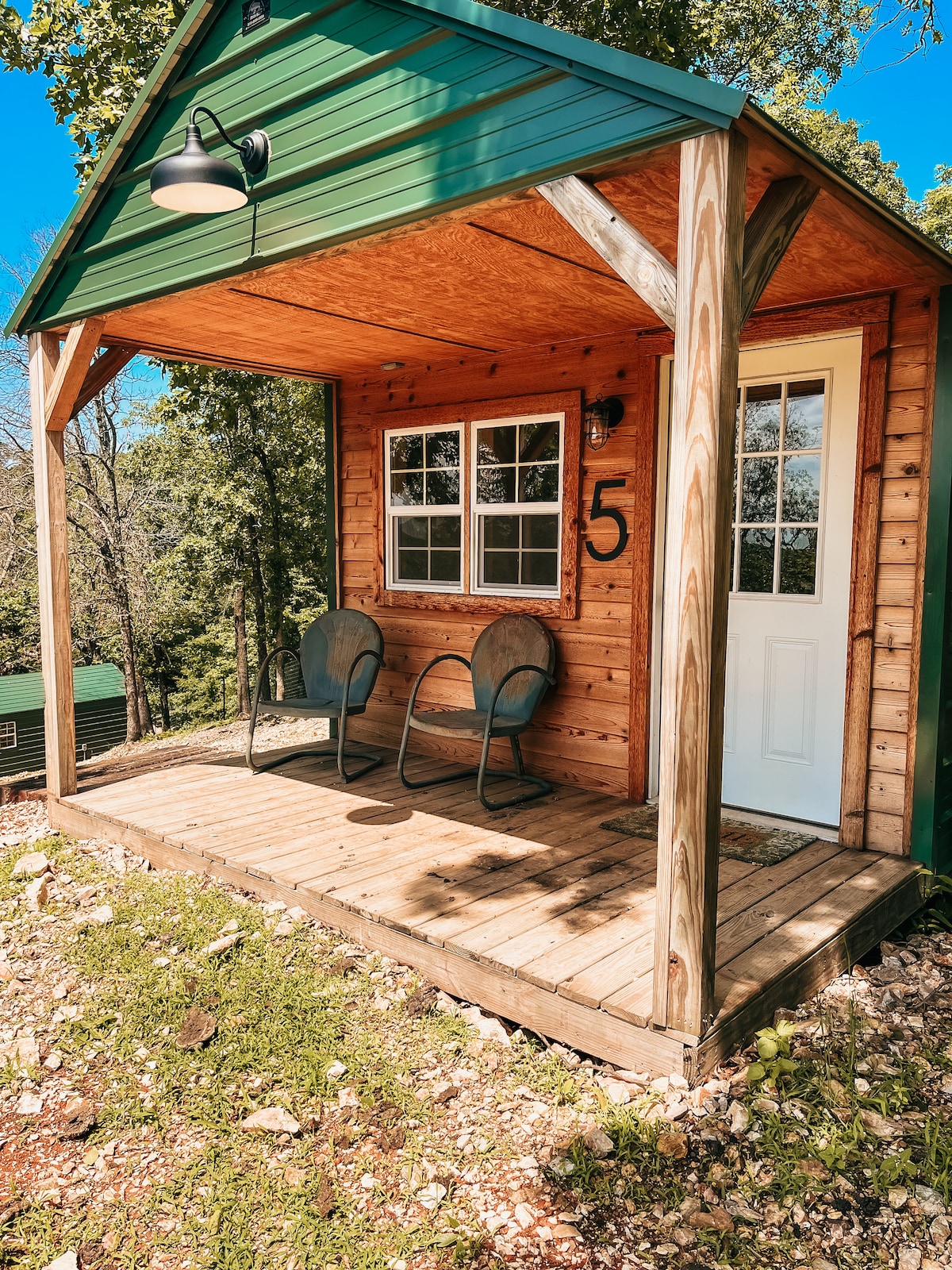 A small cabin exterior is shown, featuring a wooden deck with two chairs positioned in front. The door and window are framed with light-colored wood, and a house number is displayed on the wall. Surrounding greenery is visible, enhancing the natural setting.
