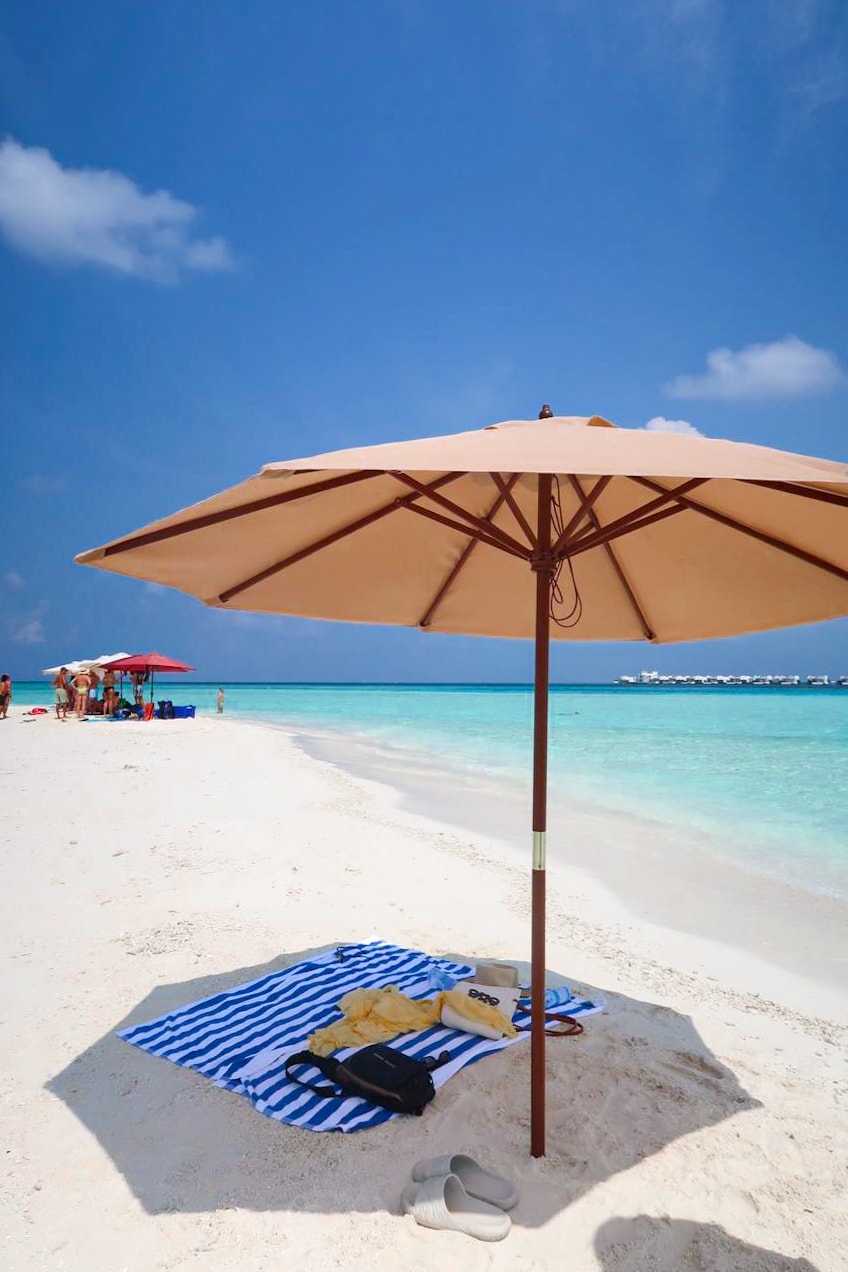 A sandy beach is depicted under a clear blue sky. A large umbrella provides shade over a striped beach blanket, accompanied by a pair of sandals. Colorful umbrellas are visible in the distance, along with calm turquoise waters lapping at the shore.