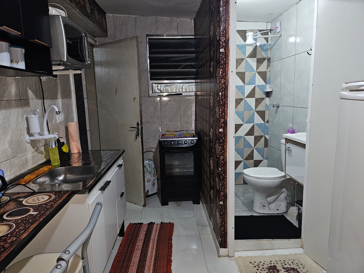 A compact kitchen and bathroom area are visible, featuring tiled walls with geometric patterns. The kitchen is equipped with a stovetop, refrigerator, and sink, while the bathroom includes a shower and toilet. A patterned rug adds a touch of detail to the space.