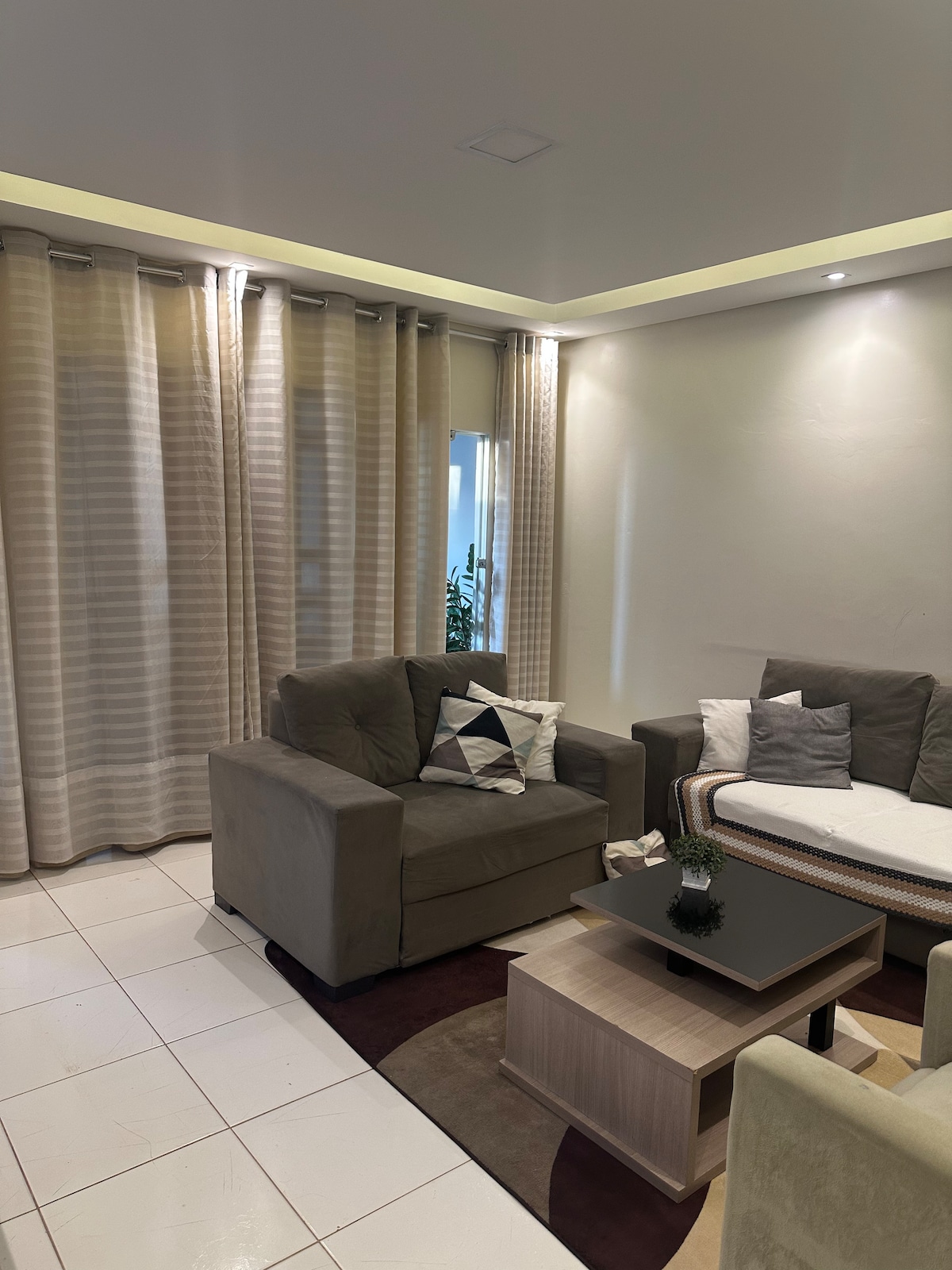 A comfortable living area is presented with two armchairs and a sofa, adorned with cushions. A coffee table sits at the center, resting on a warm-toned area rug, while sheer curtains frame the windows, allowing natural light to softly illuminate the space.