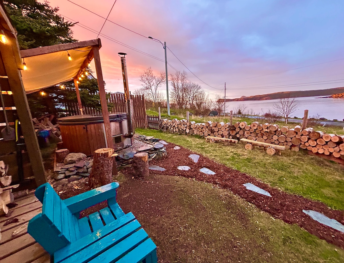 The outdoor space features a wood-fired hot tub surrounded by neatly stacked firewood and a lush grassy area. Two blue chairs are positioned on a stone pathway leading towards the water, with a backdrop of a pastel-colored sky at sunset and a fence marking the property line.