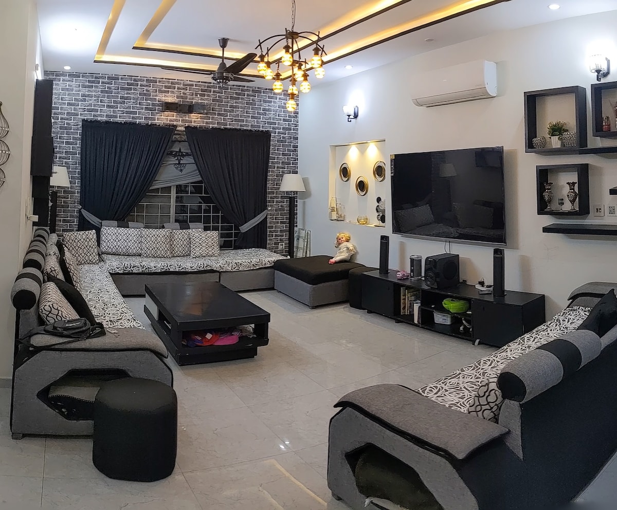A spacious TV lounge is featured, showcasing a large black coffee table at the center. Surrounding seating consists of multiple grey couches with patterned cushions. A black ottoman is positioned nearby, while an 80-inch TV is mounted on the wall, accompanied by stylish lighting fixtures.