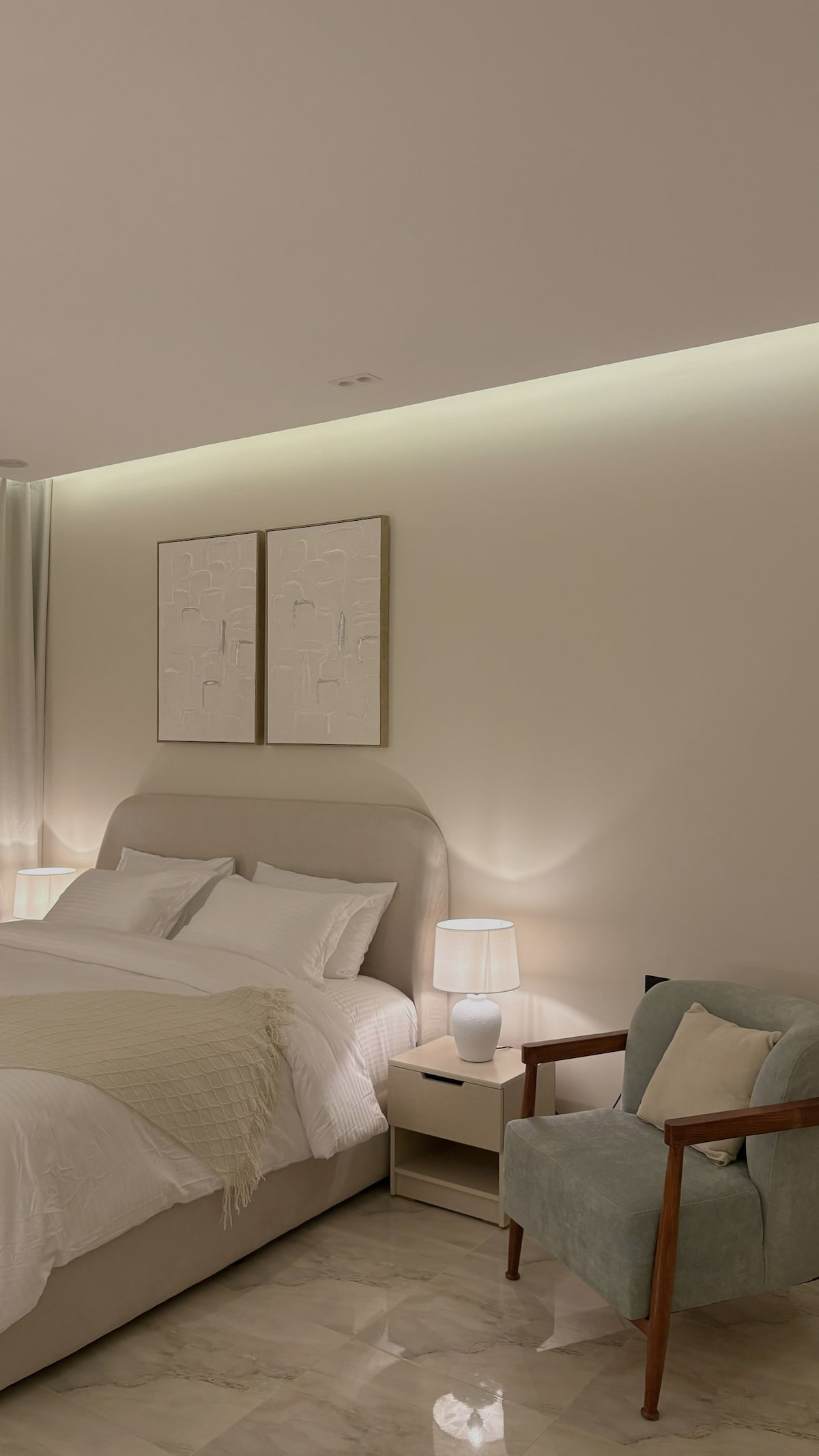 A spacious bedroom features a large, comfortable bed with neatly arranged pillows. Soft lighting is provided by bedside lamps on either side. A light-colored armchair is positioned next to a small side table. Two framed artworks adorn the wall above the bed, contributing to the serene atmosphere.