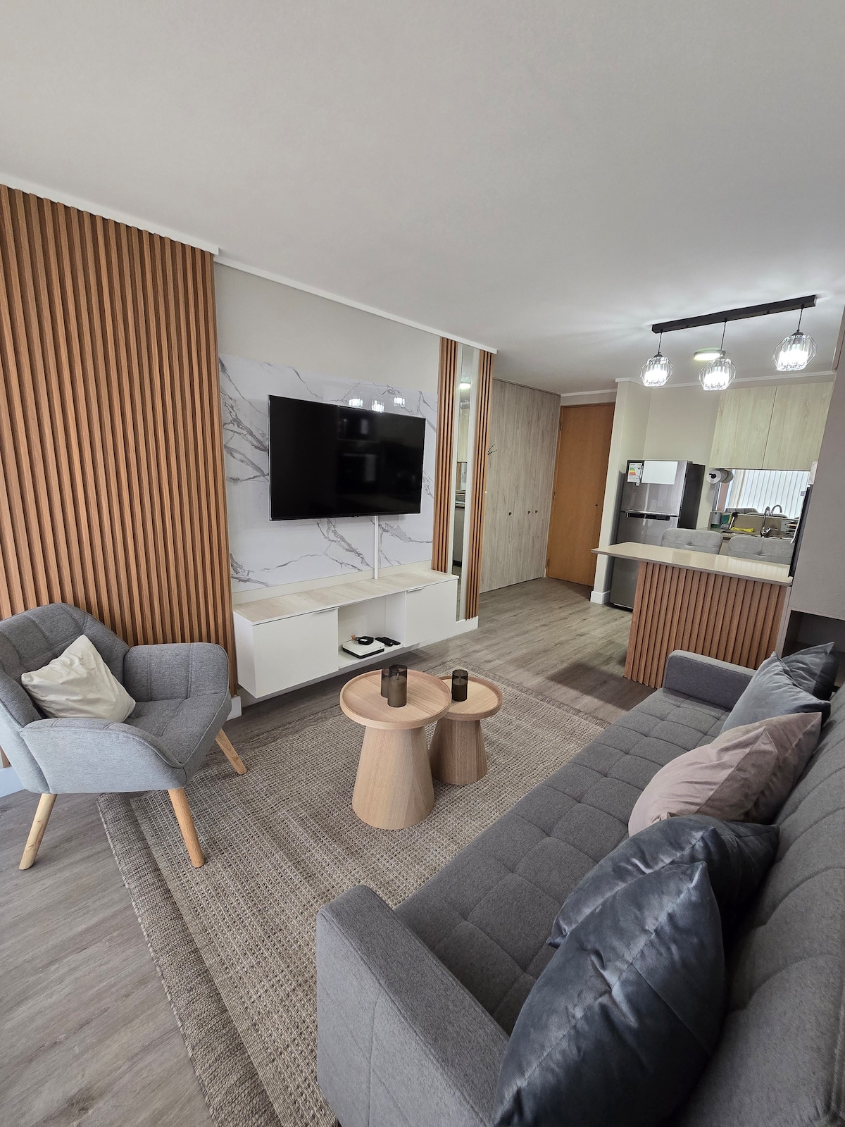 A modern living area is presented with a comfortable gray sofa and a gray armchair. A wall-mounted TV is positioned on a sleek white console beneath warm wood paneling. Light-colored flooring complements the cozy atmosphere, with a woven rug adding texture.