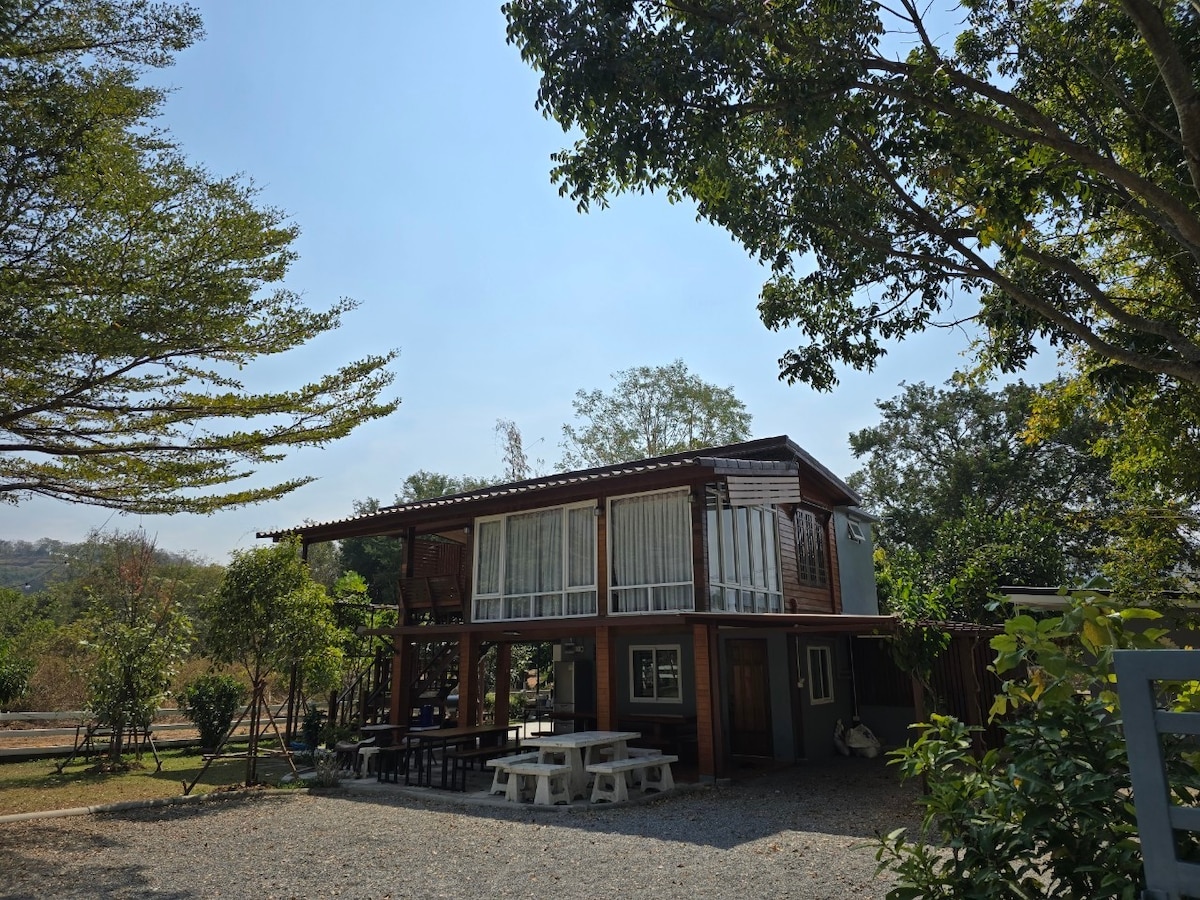 A two-story structure made of wood and glass is surrounded by lush greenery. Outdoor seating is available beneath a covered area, providing ample space for relaxation. The building features large windows that allow natural light to brighten the interiors.