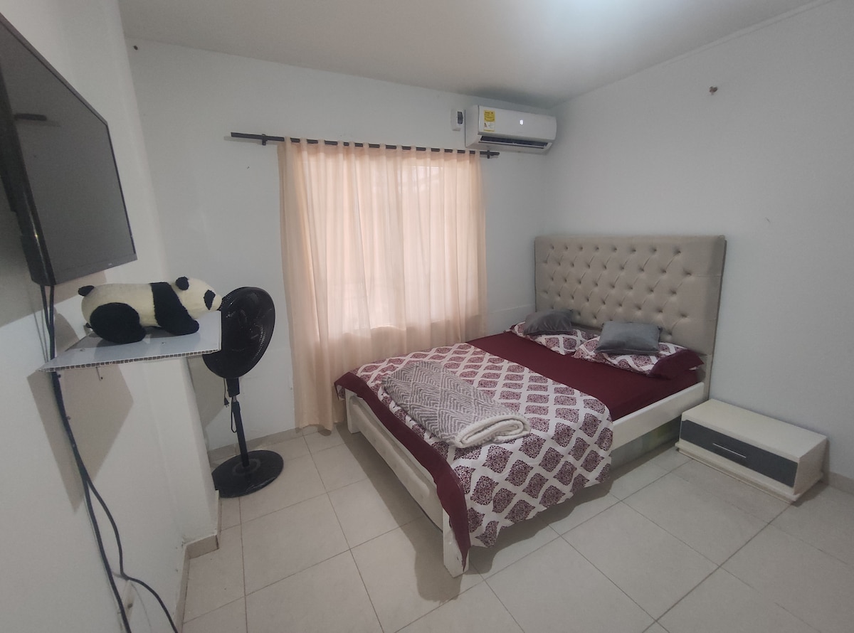 A well-lit sleeping area features a spacious bed with a patterned maroon and white bedspread. A flat-screen television is mounted on the wall, while a standing fan and a white bedside table are positioned nearby. Soft curtains filter natural light from the window.