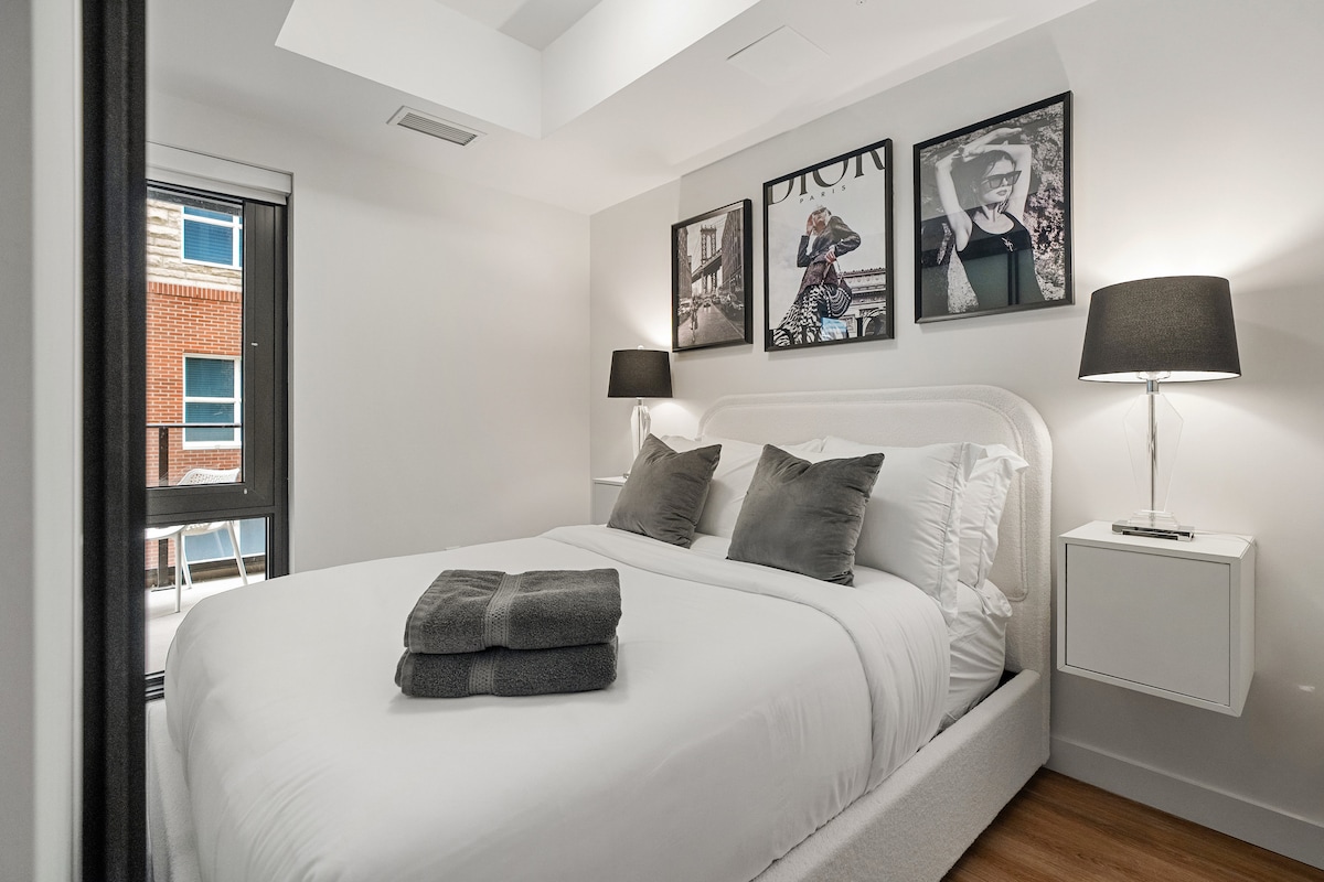 A modern bedroom features a spacious bed with crisp white linens and decorative pillows. Two bedside lamps provide soft lighting, and framed art pieces adorn the walls. A window offers a view of the exterior, enhancing the room's ambiance.