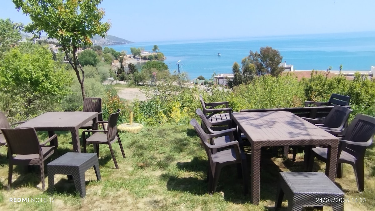 A grassy outdoor area is filled with several dining tables and chairs, positioned to overlook a view of the sea. The serene blue water complements the greenery, creating a peaceful atmosphere. Surrounding trees and plants enhance the natural setting.